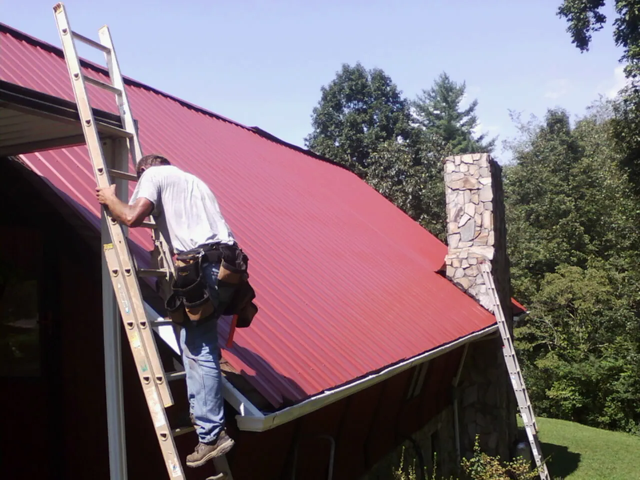 Roofer on a ladder beside a red metal roof and stone chimney — Soffit & Fascia Repair by Roofing Brothers in Sturgis